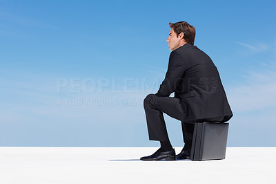 Thoughtful business man sitting on briefcase against blue sky