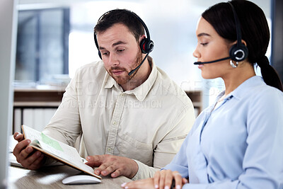 Buy stock photo Shot of two young call centre agents sitting in the office together and looking at a notebook