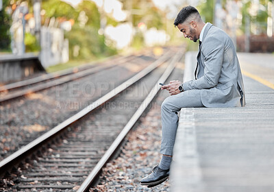 Buy stock photo Shot of a young businessman using a cellphone while sitting alongside a railway track during his commute