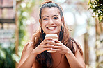 Happy young mixed race woman enjoying a cup of coffee on a break at a cafe in the city. One female only drinking a warm beverage in a disposable takeaway paper cup while relaxing outside a restaurant