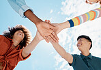 A low close up view of a group of diverse young friends joining hands in a huddle while smiling with a blue sky in the background on a sunny day. Mixed race female with a cool afro hairstyle and her asian man friend showing unity with a group of friends