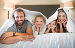 Young caucasian family lying under a blanket in a bed together at home. Cute little siblings relaxing in bed with their mother and father in a bedroom