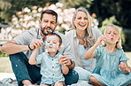 Smiling caucasian family blowing soap bubbles for fun while relaxing together in the park or garden outside on a sunny day. Loving parents bonding with playful kids enjoying a carefree happy childhood