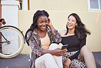 Two multiethnic female friends sitting on sidewalk and laughing while browsing social media. Best friends checking smartphone content while sitting on street