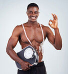Portrait of a smiling African American fitness model posing topless with a scale and measuring tape. Happy black male athlete isolated on grey copyspace showing the OK symbol for a balanced diet