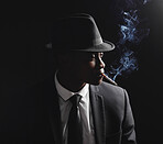 African man wearing a formal suit and hat while smoking against a black background. Dapper and classy young black guy with a cigarette in his mouth. Vintage hitman from the mafia looking thoughtful