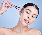 A beautiful mixed race woman posing with a makeup brush during a pamper routine. Hispanic model holding a contouring brush against a blue copyspace background