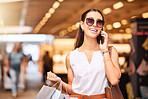 A happy young mixed race woman wearing sunglasses using a cell phone while carrying bags during a shopping spree. Young brunette woman on a call with her smartphone while enjoying shopping in a mall. A little retail therapy is never a bad idea