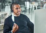 Young male call center agent smiling and wearing a headset and mask working on a computer in an office at work. Customer service, support and sales. Giving advice and helping. Answering calls
