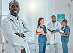 Mature african american male doctor standing with his arms crossed while working at a hospital. One expert medical professional smiling while standing at work with colleagues at a clinic