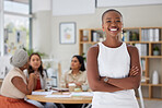 Portrait of smiling african american business woman standing with her arms folded while her colleagues sit behind her in the office. Ambitious, confident and happy black professional with arms crossed