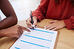 Closeup of two unknown ethnic business woman sitting and signing office contract. African american professional using hand gesture to point on paperwork. Mixed race colleague agreeing to document deal
