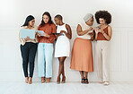 Group of five diverse young businesswomen standing against a wall in an office and using tech. Happy colleagues talking and using technology while standing in a row together at work