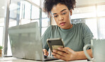 Young serious mixed race woman working a laptop and using a phone sitting at a table at home. One focused hispanic female with a curly afro planning and using social media on a cellphone alone in the lounge at home
