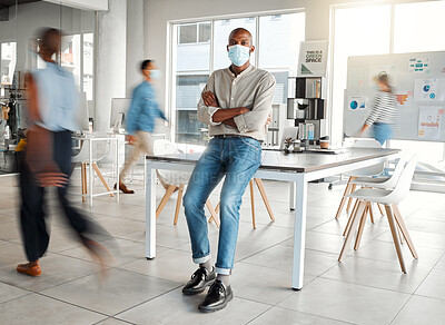 Buy stock photo Young African american businessman standing with his arms crossed and wearing a mask at work. One confident male manager wearing a mask protecting from a virus and standing in a busy office at work