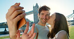 Young man taking a selfie while his girlfriend kisses his cheek on holiday in London